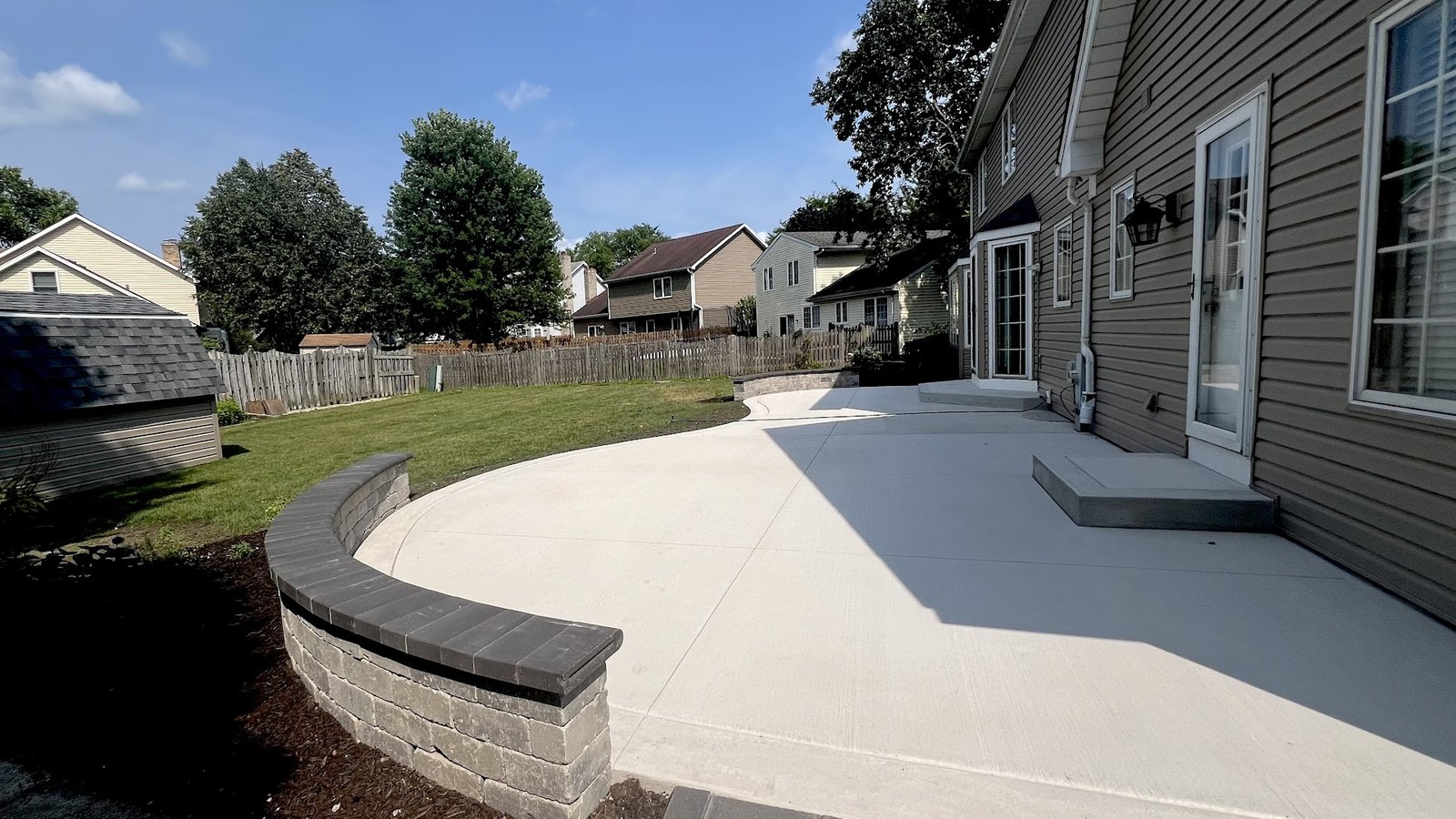 Hardscape contractors in Des Plaines: finished curved poured concrete patio with charcoal-cap retaining wall border on a Cook County backyard install
