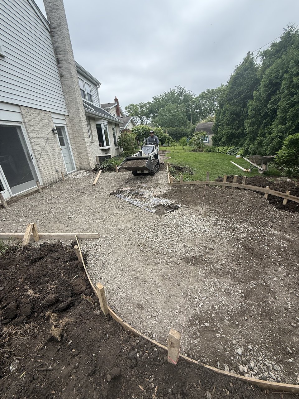 Hardscape contractors in Des Plaines: skid-steer placing crushed limestone over excavated subsoil for a curved patio formwork ahead of pour